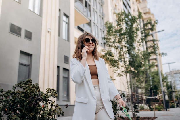 Mujer de negocios sonriente con traje blanco hablando por teléfono mientras camina por la ciudad con arquitectura moderna