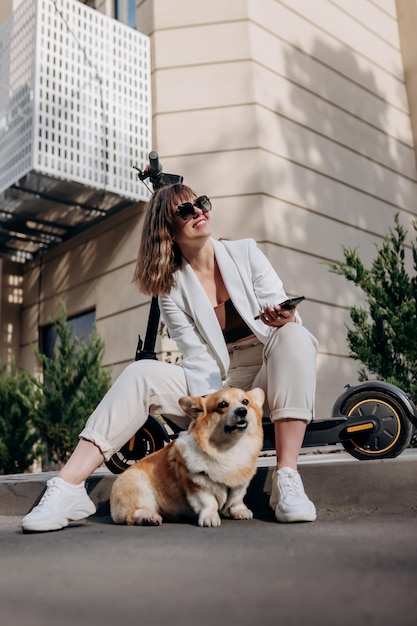 Mujer de negocios sonriente con traje blanco sentada en scooter eléctrico y usando el teléfono mientras camina con el perro Welsh Corgi Pembroke en la ciudad