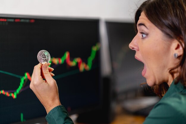 Mujer de negocios sorprendida sosteniendo y mirando una moneda ethereum. Comercio en línea, dinero virtual y concepto de criptomoneda