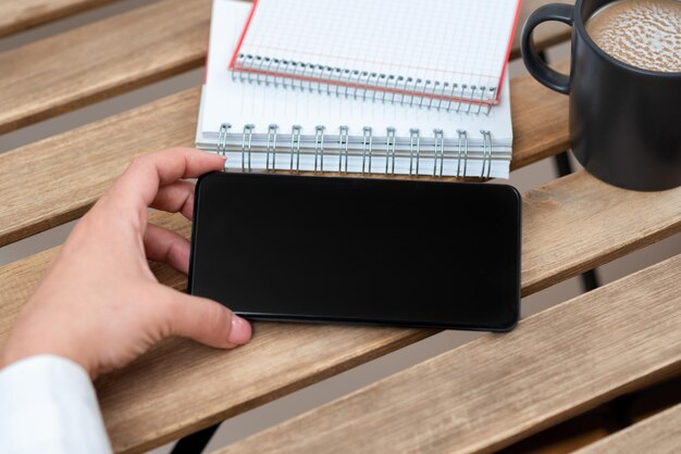 Mujer de negocios sosteniendo un teléfono celular con un mensaje importante en la mesa con café y un cuaderno Mujer sentada presentando información crucial en la pantalla del teléfono