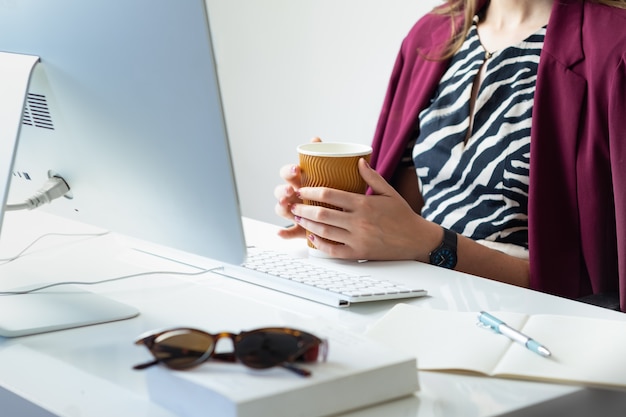 Mujer de negocios con taza de café en el escritorio de oficina minimalista. Persona de sexo femenino joven delante de la computadora de escritorio en el lugar de trabajo moderno