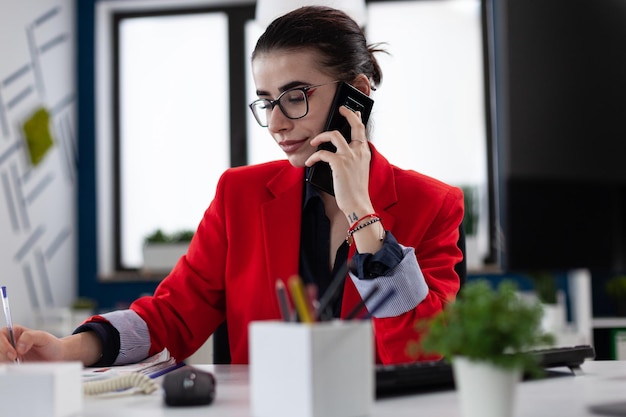 Mujer de negocios tomando notas en una llamada de teléfono inteligente sentada en el escritorio. Empresario con gafas trabajando en la oficina de inicio. Propietario de una pequeña empresa con chaqueta roja escribiendo en papel en un entorno de inicio.