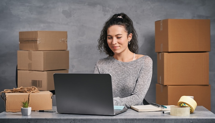 Foto mujer de negocios trabajando desde casa con un portátil y un bloc de notas preparando cajas con productos para la entrega