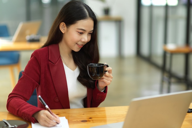 Mujer de negocios trabajando en la oficina. tomando notas mientras mira hacia abajo en la computadora portátil y sostiene una taza de café. Empresaria ocupada