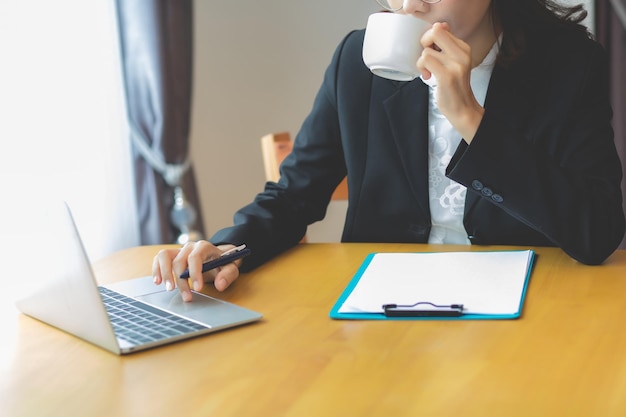 Una mujer de negocios con traje negro está sentada en un escritorio escribiendo una laptop y bebiendo café en su oficina