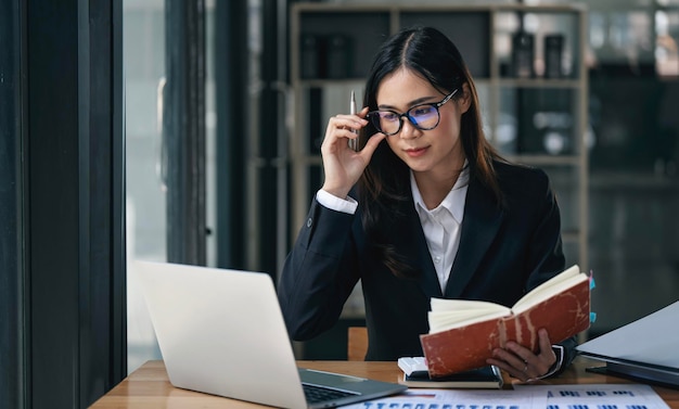 Mujer de negocios en traje trabajando en una computadora portátil en la oficina con las manos en las gafas y sosteniendo un cuaderno