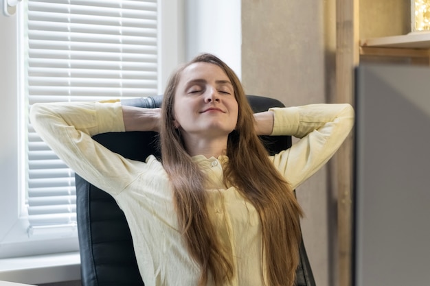 Mujer de negocios tranquila y feliz en su oficina se sienta en un sillón de cuero con las manos detrás de la cabeza.