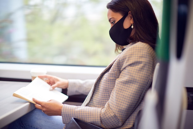Mujer de negocios en el tren relajándose y leyendo un libro con mascarilla facial PPE durante la pandemia de salud