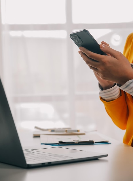 Foto mujer de negocios usando teléfono móvil mientras trabaja en una computadora portátil navegando por internet buscando datos comerciales en una oficina moderna mujer de negocios asiática en línea trabajando en la computadora en el lugar de trabajo