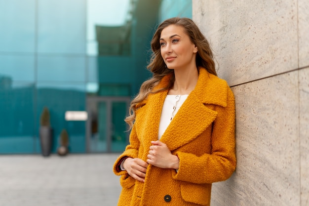 Mujer de negocios vestida con abrigo amarillo de pie al aire libre superficie del edificio corporativo Persona de negocios femenina caucásica en la calle de la ciudad cerca del edificio de oficinas con ventanas Elegante empresaria