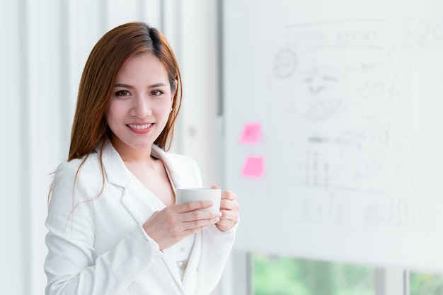 La mujer de negocios en el vestido blanco está sosteniendo la taza de café en la sala de reunión