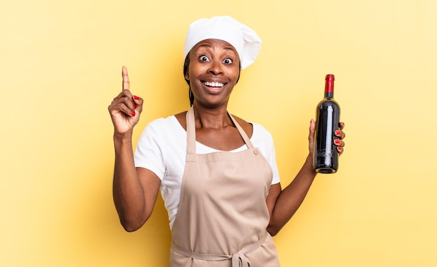 Mujer negra afro chef sintiéndose como un genio feliz y emocionado después de darse cuenta de una idea, levantando alegremente el dedo, ¡eureka !. concepto de botella de vino