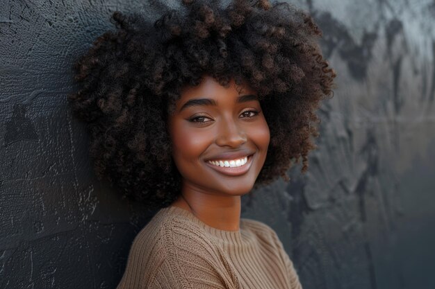 Una mujer negra alegre con un afro voluminoso sonríe brillantemente mirando a la cámara apoyándose en una pared negra de la ciudad concepto de actitud positiva