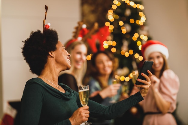 Foto mujer negra alegre divirtiéndose y haciendo videollamadas con su smartphone mientras celebra el año nuevo en casa con sus amigas.