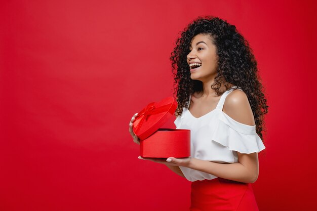 Mujer negra bastante alegre que abre el regalo de San Valentín en forma de corazón aislado en rojo