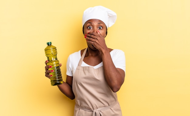 Mujer negra chef afro cubriéndose la boca con las manos con una expresión de asombro y sorpresa, manteniendo un secreto o diciendo uy. concepto de aceite de oliva