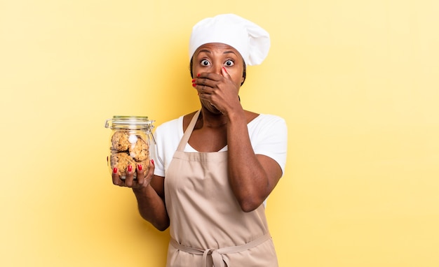 Mujer negra chef afro cubriéndose la boca con las manos con una expresión de asombro y sorpresa, manteniendo un secreto o diciendo uy. concepto de cookies