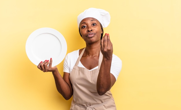 Mujer negra chef afro haciendo capice o gesto de dinero, ¡diciéndote que pagues tus deudas !. concepto de plato vacío