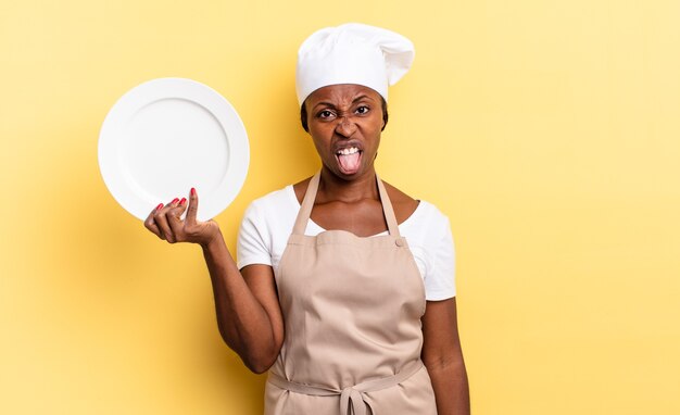 Mujer negra chef afro que se siente disgustada e irritada, sacando la lengua, disgustando algo desagradable y asqueroso. concepto de plato vacío