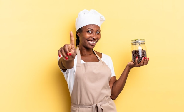 Mujer negra chef afro sonriendo con orgullo y confianza haciendo la pose número uno triunfalmente, sintiéndose como una líder. concepto de granos de cafe