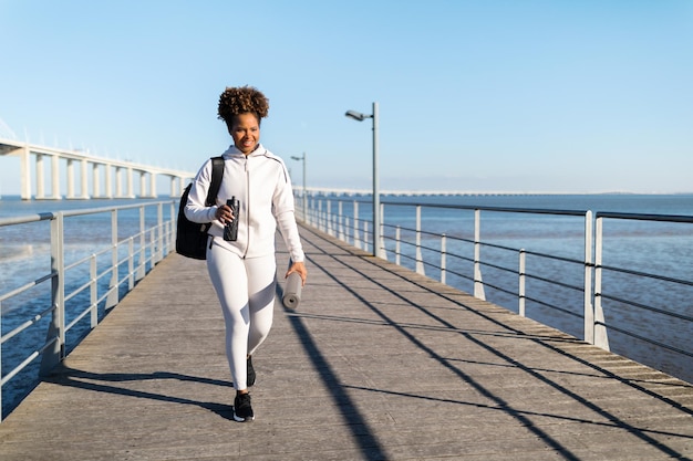 Mujer negra con colchoneta de yoga y botella de agua en las manos caminando al aire libre