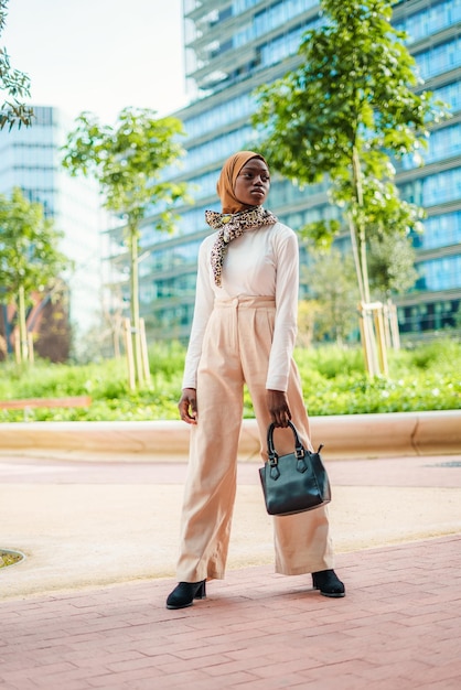Mujer negra confiada en traje de moda y bolso de mano hijab