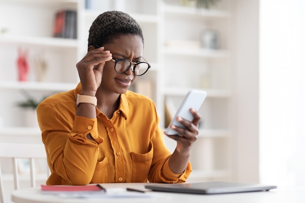 Mujer negra confundida mirando la pantalla del teléfono inteligente tocando gafas