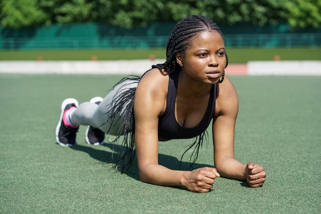 Una mujer negra determinada hace ejercicio de tablones en el suelo de césped verde del estadio de la ciudad contra el parque