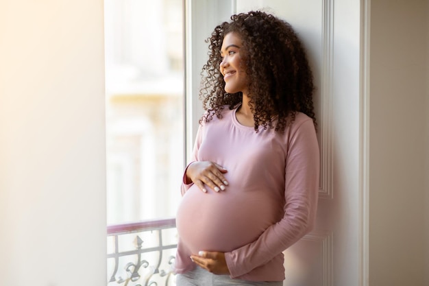Mujer negra embarazada acunando suavemente el vientre mientras está de pie junto a la ventana en casa