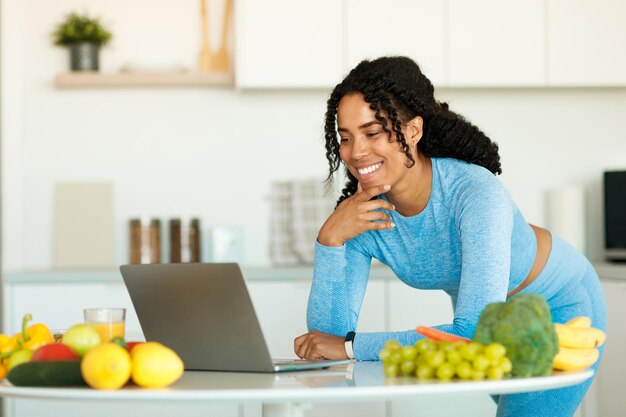 Una mujer negra emocionada usando una laptop en la cocina buscando recetas saludables en línea mientras cocina el almuerzo después del entrenamiento doméstico