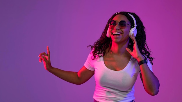Mujer negra feliz con auriculares y gafas de sol con estilo posando en luz de neón