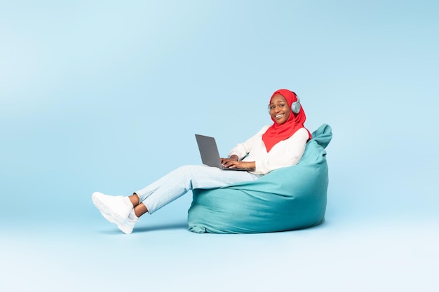 Mujer negra feliz en hiyab usando una computadora portátil y usando auriculares sentado en una silla de frijoles espacio de copia de fondo azul