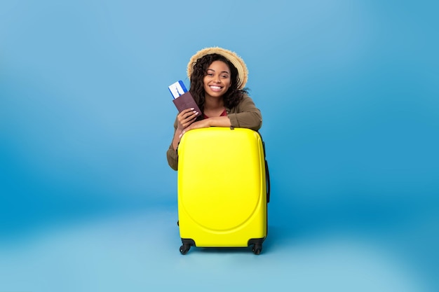 Mujer negra feliz con ropa informal sentada detrás de una maleta con pasaporte y billetes de avión en marcha