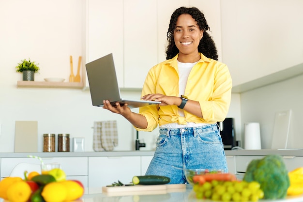 Mujer negra feliz sosteniendo y usando una computadora portátil buscando nuevas recetas cocinando comida saludable en una cocina moderna