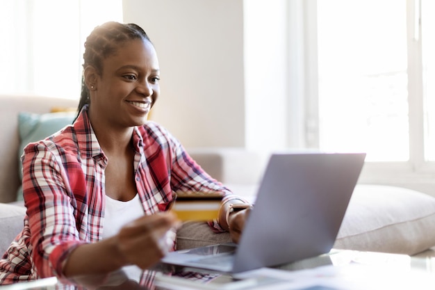 Mujer negra feliz usando laptop y tarjeta bancaria en casa