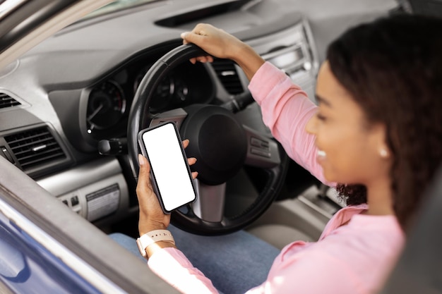 Mujer negra feliz usando un teléfono inteligente con una pantalla vacía conduciendo un automóvil