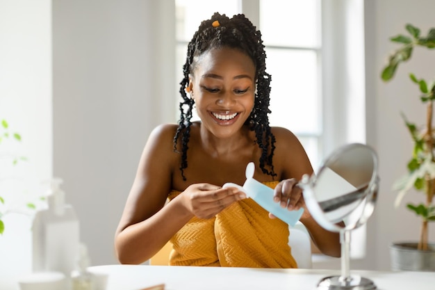 Mujer negra feliz vertiendo agua micelar en una almohadilla de algodón haciendo rutina de belleza en casa limpiando la cara