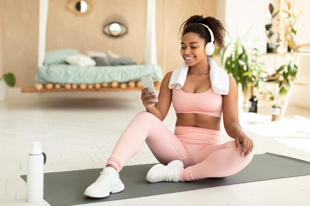 Mujer negra en forma positiva con toalla en los hombros y auriculares inalámbricos sentados en una alfombra de fitness y escuchando música