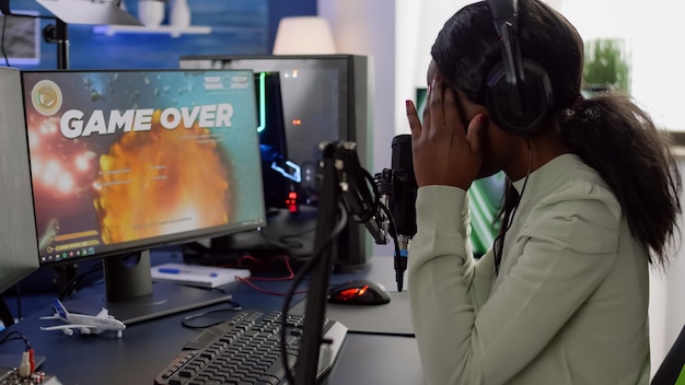 Mujer negra furiosa gamer con auriculares usando chat de transmisión perdiendo el juego jugando en un poderoso juego de disparos de computadora RGB