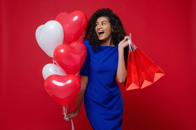 Mujer negra con globos de colores en forma de corazón y bolsas de compras en la pared roja