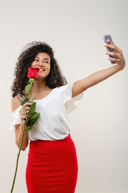 Mujer negra haciendo selfie con una rosa con falda roja y blusa blanca aislada