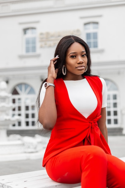 La mujer negra joven de moda moderna en traje rojo elegante hermoso está descansando en el banco de madera en la ciudad. Modelo de moda glamorosa chica africana de moda en traje de negocios vintage posa en la calle.