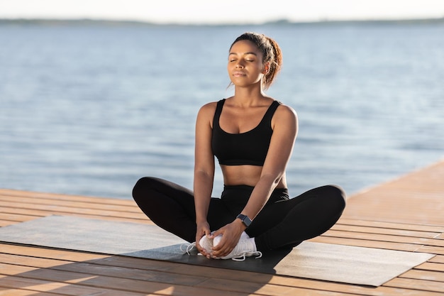 Mujer negra joven pacífica que medita al lado del lago de la ciudad