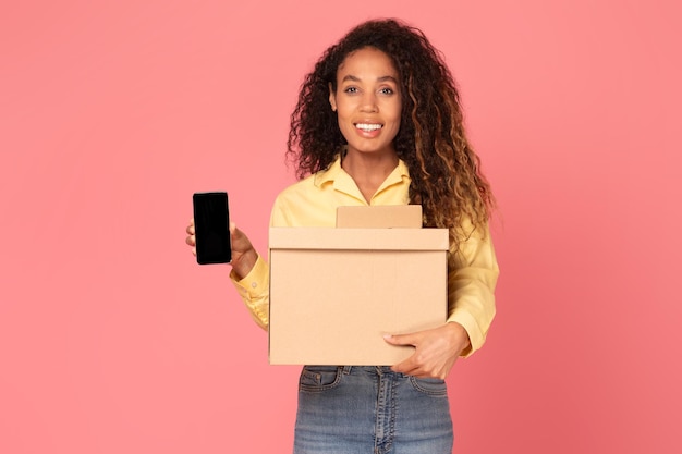 Mujer negra joven con paquetes de caja y pantalla de teléfono en blanco sobre fondo rosa