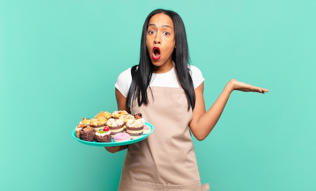 Mujer negra joven que parece sorprendida y consternada, con la mandíbula caída sosteniendo un objeto con una mano abierta en el costado. concepto de chef de panadería