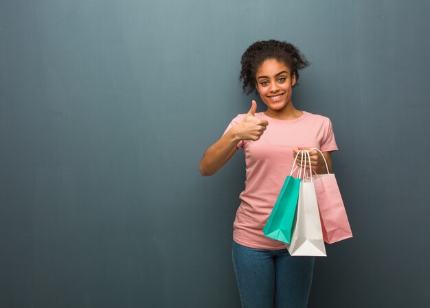 Mujer negra joven que sonríe y que levanta el pulgar para arriba. Ella está sosteniendo una bolsa de compras.