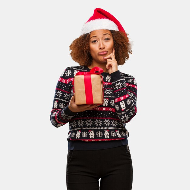 Mujer negra joven que sostiene un regalo en el día de la Navidad que duda y confusa