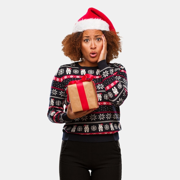 Mujer negra joven que sostiene un regalo en el día de Navidad sorprendida y chocada