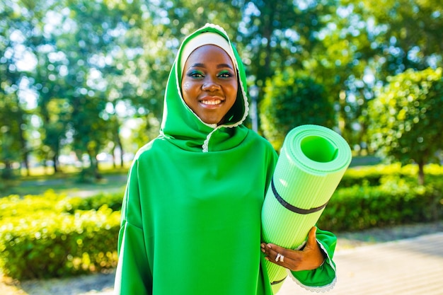 Mujer negra latina en hiyab verde con maquillaje brillante y colchoneta de yoga holg de nariz penetrante al aire libre en el parque de verano