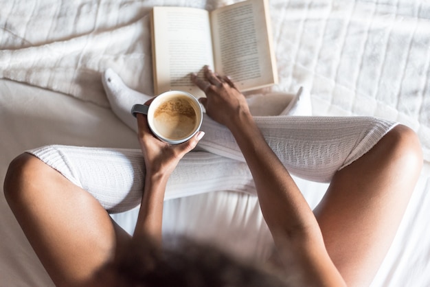 Mujer negra leyendo un libro y tomando café en la cama con calcetines